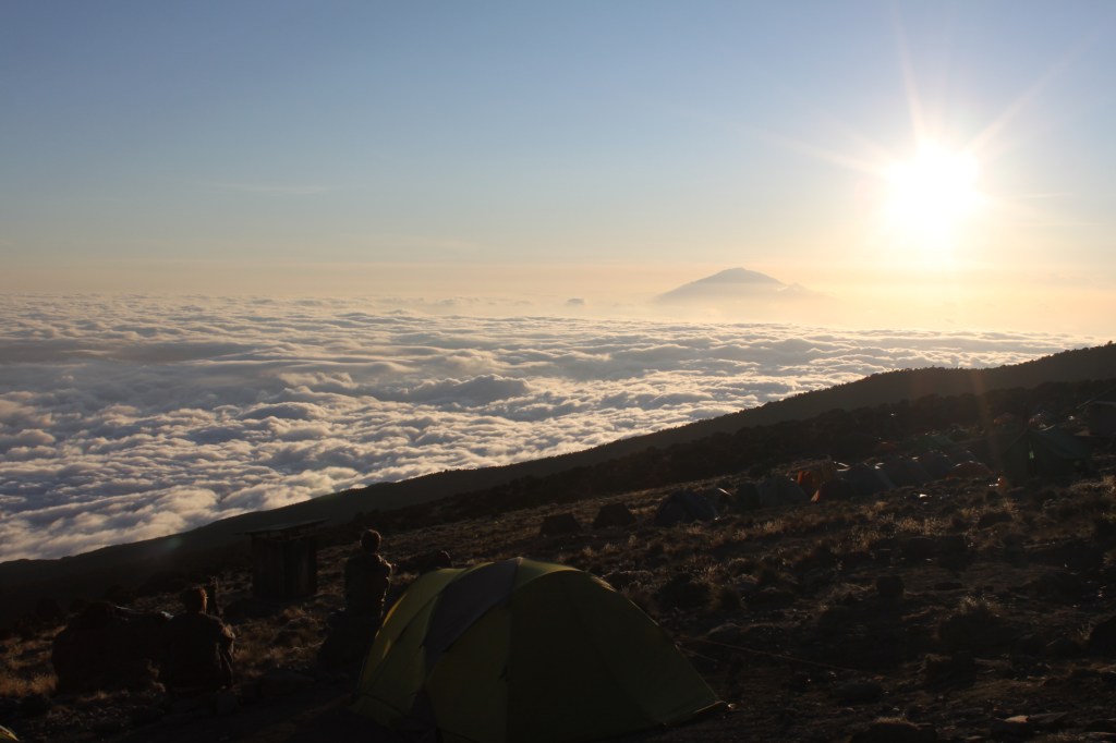 The sun starts to set over Mt Kilimanjaro with onlookers sitting on the side of the mountain overlooking a sky full of clouds.