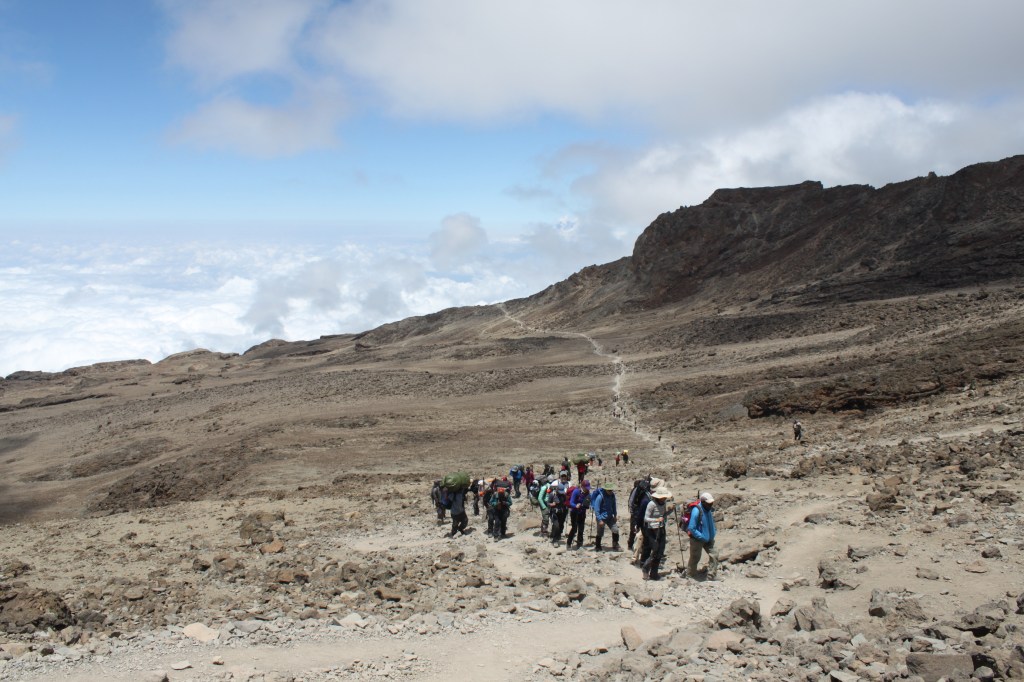 A group of trekkers walks a long, rocky open path on a trek to the top of Mount Kilimanjaro.