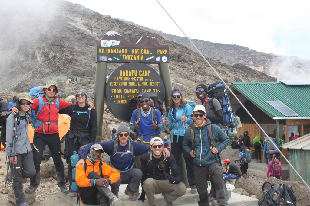 A group of 10 smiling trekkers standing at the Barafu Camp sign in Kilimanjaro National Park.